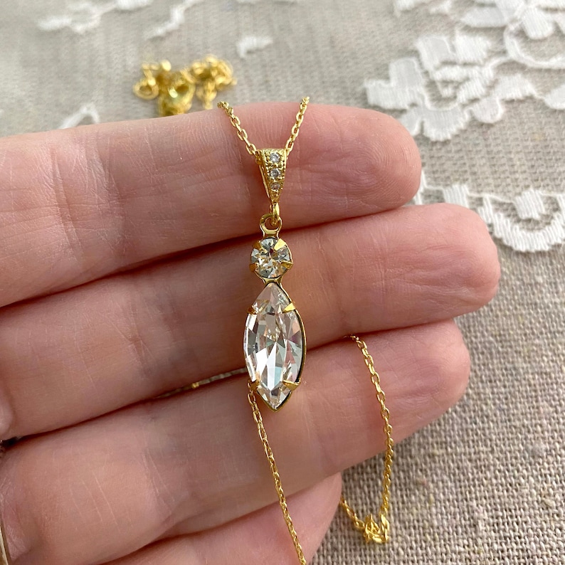 May include: A gold-colored necklace with a large, clear marquise-cut crystal pendant. The pendant is suspended from a delicate chain and features a smaller round crystal above it. The necklace is held against a neutral background.