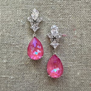 May include: A pair of silver-toned drop earrings. Each earring features a cluster of clear, sparkling crystals at the top, with a large, pear-shaped, pink crystal pendant. The pink crystals have an iridescent quality, reflecting multiple colors.