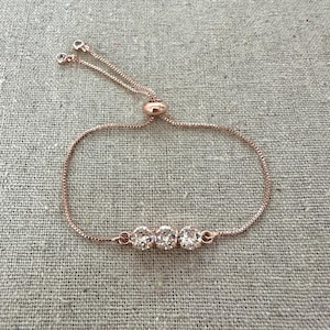 May include: Rose gold adjustable bracelet featuring three clear, round cubic zirconia stones. The bracelet has a delicate chain and a sliding bead closure for a customizable fit. The stones are set in a row, adding a touch of sparkle.