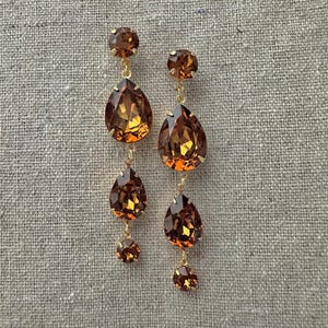 May include: A pair of drop earrings featuring gold-tone metal settings and faceted amber-colored crystals. Each earring consists of a round top stone, a large pear-shaped center stone, a smaller pear-shaped stone, and a tiny round bottom stone.
