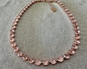 May include: A rose gold tone necklace featuring a continuous strand of round, faceted light pink crystals set in metal bezels. The necklace includes a short extender chain with a small rectangular metal tag attached at the clasp mechanism.