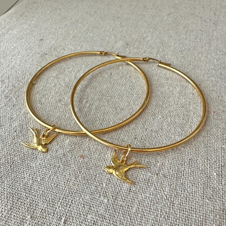 May include: A pair of gold-colored hoop earrings. Each earring features a small, detailed bird charm hanging from the hoop. The earrings are displayed on a neutral-colored, textured surface.