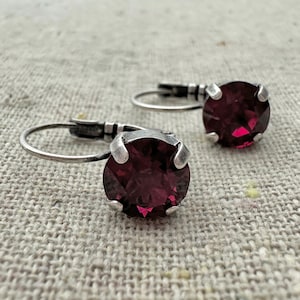 May include: A pair of leverback earrings featuring large, round, faceted, deep red gemstones set in silver-toned metal. The earrings are displayed on a textured, neutral-colored surface.