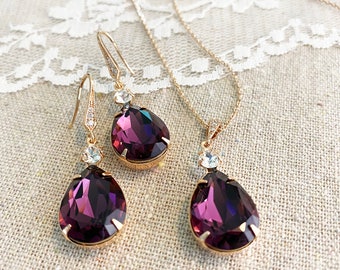 May include: A gold-toned jewelry set featuring a necklace and a pair of earrings. Each piece showcases a large, teardrop-shaped, deep purple crystal pendant, accented with smaller clear crystals. The set is displayed on a neutral-toned fabric.