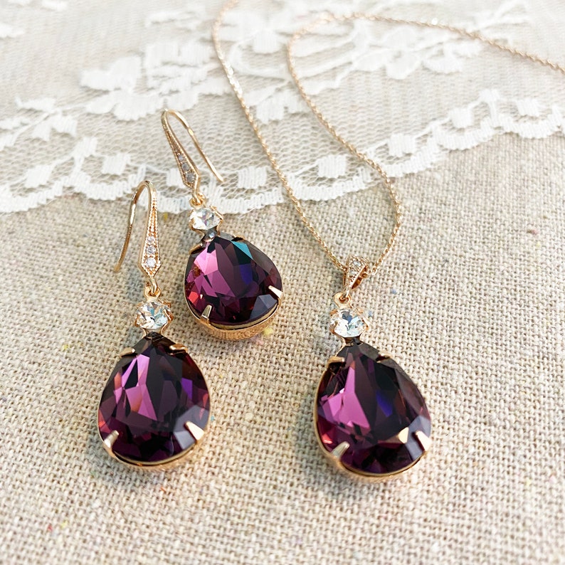 May include: A gold-toned jewelry set featuring a necklace and a pair of earrings. Each piece showcases a large, teardrop-shaped, deep purple crystal pendant, accented with smaller clear crystals. The set is displayed on a neutral-toned fabric.