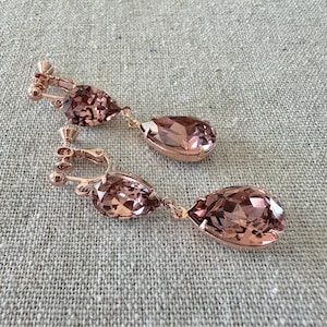 May include: A pair of rose-gold clip-on earrings with large, faceted, teardrop-shaped pink crystals. The earrings have a delicate design with a clip-on mechanism for non-pierced ears. The crystals catch the light, creating a sparkling effect.