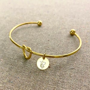 May include: A gold-coloured bangle bracelet featuring a knot design and a small, round charm. The charm is engraved with the letter "B". The bracelet has a simple, elegant design.