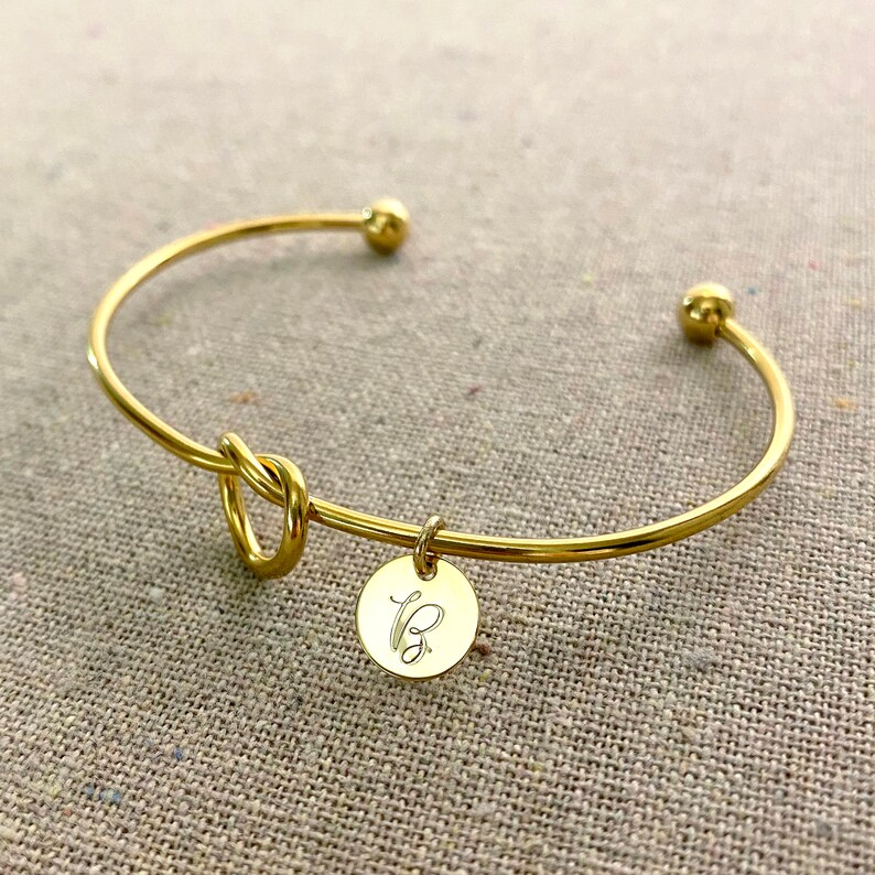 May include: A gold-coloured bangle bracelet featuring a knot design and a small, round charm. The charm is engraved with the letter "B". The bracelet has a simple, elegant design.