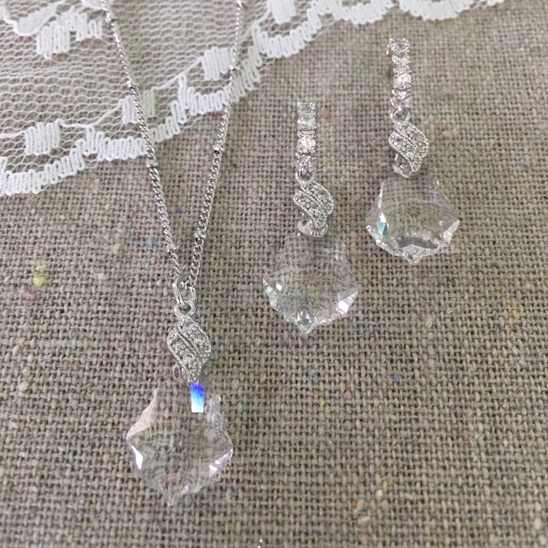 May include: A silver necklace and a pair of matching earrings. The necklace has a delicate chain with a clear, faceted crystal pendant. The earrings have a similar design, with clear crystals and silver detailing. The jewellery set is displayed on a textured, neutral-toned surface.