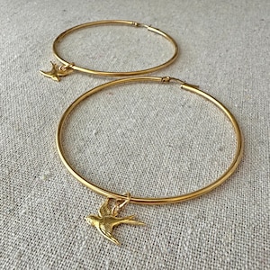 May include: Two gold-colored hoop earrings with a bird charm. The earrings are circular and have a shiny finish. Each earring has a small, detailed bird pendant hanging from it. The background is a neutral, textured fabric.