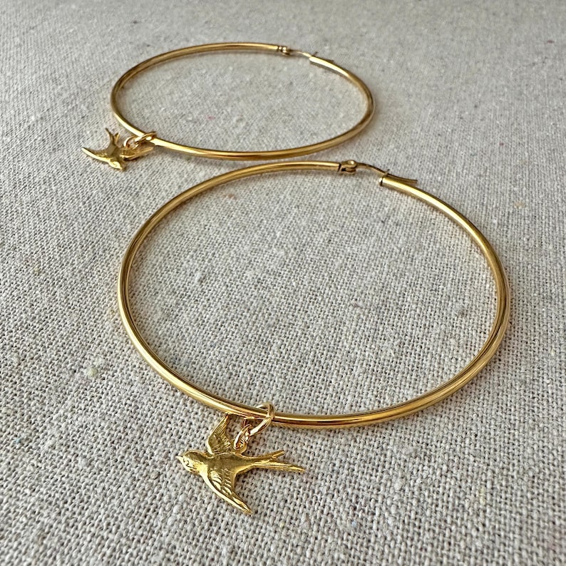 May include: Two gold-colored hoop earrings with a bird charm. The earrings are circular and have a shiny finish. Each earring has a small, detailed bird pendant hanging from it. The background is a neutral, textured fabric.