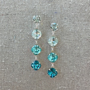 May include: A pair of dangling earrings with a series of round, faceted, aquamarine-colored crystals. The earrings have a silver-colored chain connecting the crystals, with the largest crystal at the bottom. The background is a neutral gray fabric.