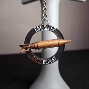 Peut inclure: Un porte-clés avec un pendentif en forme de bombe de couleur bronze à l'intérieur d'un anneau noir. L'anneau porte les mots "EAT, SLEEP" au-dessus de la bombe et "RAID, REPEAT" en dessous. Le porte-clés est attaché à une chaîne argentée.