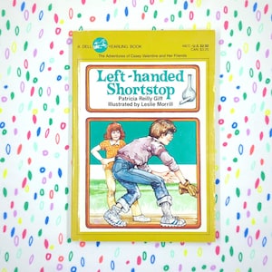 May include: A vintage paperback book titled "Left-handed Shortstop" by Patricia Reilly Giff, illustrated by Leslie Morrill. The cover features a baseball scene with two children in a yellow and green frame. The background is a white surface with colorful, abstract shapes.