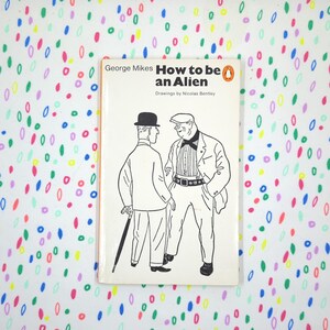 May include: A vintage paperback book titled "How to be an Alien" by George Mikes, with illustrations by Nicolas Bentley. The cover features a black and white illustration of two men. The background is a white surface with colorful, abstract shapes.