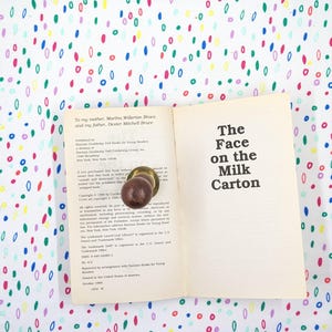 May include: An open book titled "The Face on the Milk Carton" with a brown doorknob on the pages. The book is on a white surface covered in colorful, oval-shaped patterns. The book's title is in bold, black text.