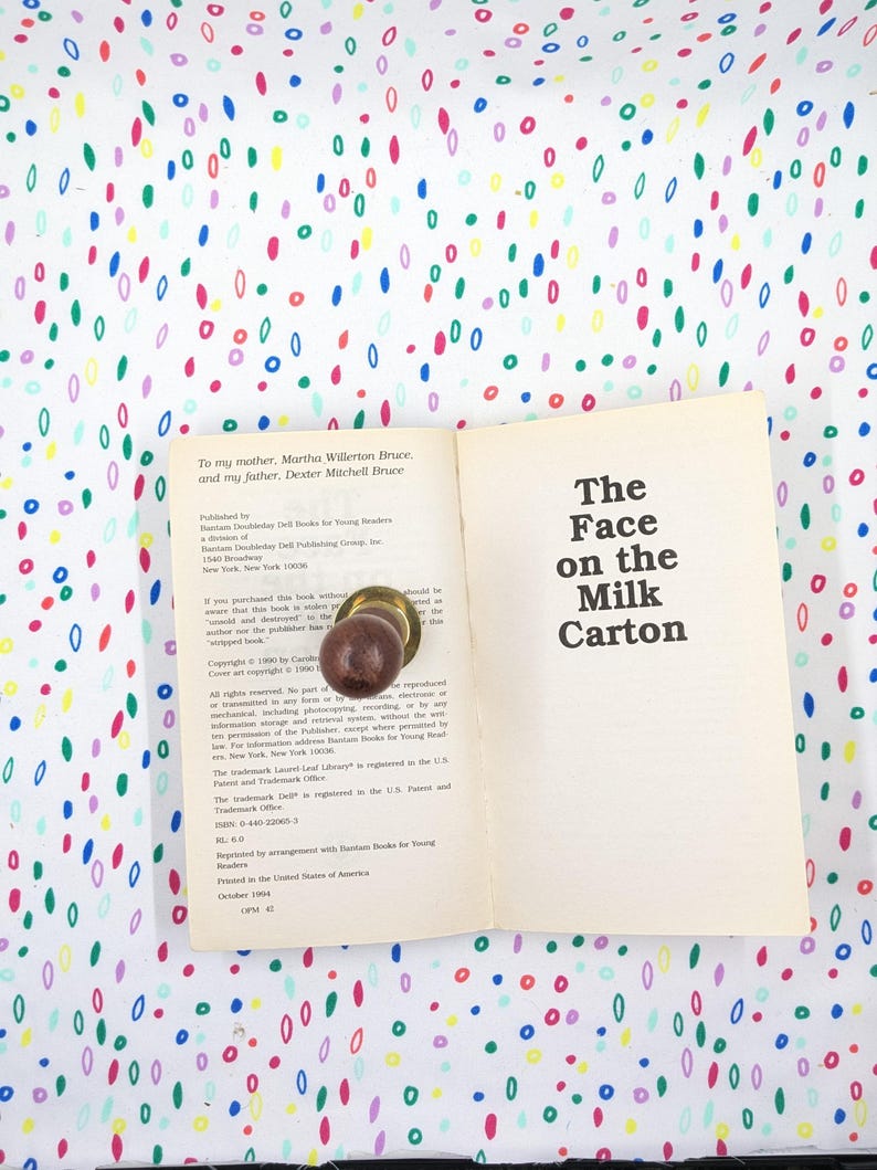 May include: An open book titled "The Face on the Milk Carton" with a brown doorknob on the pages. The book is on a white surface covered in colorful, oval-shaped patterns. The book's title is in bold, black text.