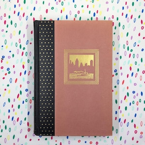 May include: A pink hardcover book with a black spine and gold accents. The front cover features a gold-framed illustration of a city skyline.
