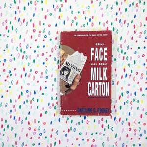 May include: A hardcover book titled "The Face on the Milk Carton" by Caroline B. Cooney. The book has a red cover with white text and a missing person's flyer. The background is a white fabric with colorful oval shapes.