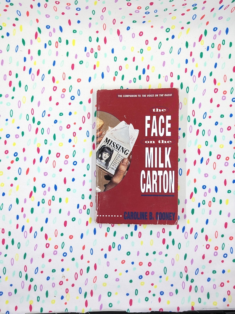 May include: A hardcover book titled "The Face on the Milk Carton" by Caroline B. Cooney. The book has a red cover with white text and a missing person's flyer. The background is a white fabric with colorful oval shapes.