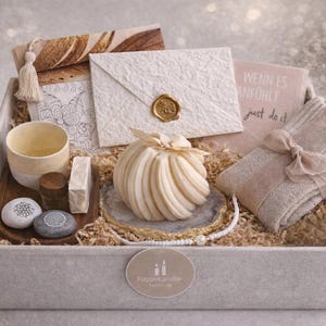 Könnte beinhalten: Eine Geschenkbox mit Pflegeprodukten. Sie enthält eine Kerze, Seife, Steine, eine Tasse, ein Notizbuch mit Text und einen Umschlag. Die Box ist mit Papier ausgekleidet und hat ein neutrales Farbschema.