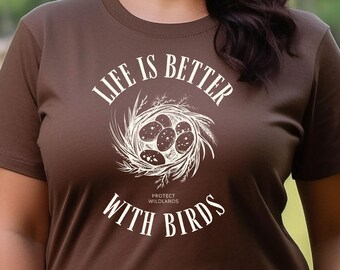 Op de afbeelding: Een bruin T-shirt met een witte afbeelding waarop staat "Life is better with birds". De afbeelding toont een vogelnest met eieren en de tekst "Protect Wildlands".