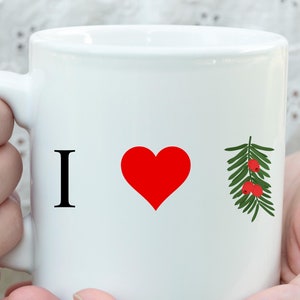 May include: White ceramic mug with the text "I ❤️" followed by a green sprig with red berries. The mug is held by a person.