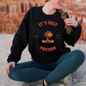 以下が含まれることがあります： オレンジ色のグラフィックが施された黒いスウェットシャツ。グラフィックには「It's Just Friction」という文字と、火を起こすために棒をこすり合わせているスタイリッシュな手が描かれている。「Est. 30,000 BC」という文字がグラフィックの下にある。