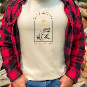 May include: A cream-colored t-shirt with a graphic of a frog in a sun-shaped arch. The text "May the Frogs Be With You" is printed on the shirt.