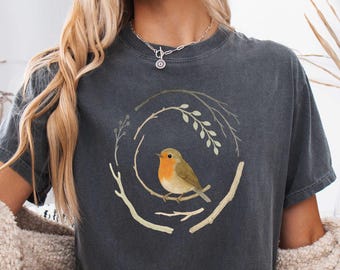 May include: Dark gray t-shirt featuring a watercolor illustration of a robin perched on a branch within a circular nest design. The bird has orange and brown feathers. The shirt is worn with denim shorts and a cream-colored cardigan.