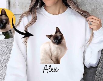 May include: White sweatshirt featuring a Siamese cat portrait with the name "Alex" in a script font. The cat has a cream-colored body, dark brown face, ears, paws, and tail, and blue eyes. A smaller cat portrait is in a yellow circle.