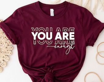 May include: A maroon t-shirt with the text "You Are You Are Enough" in white.