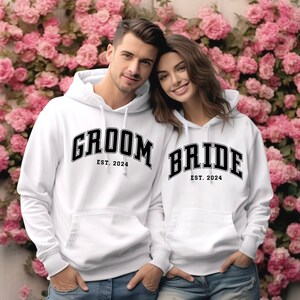 May include: A couple wearing white hoodies with the words "Groom Est. 2024" and "Bride Est. 2024" printed on them. They are standing in front of a wall of pink flowers.