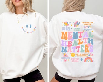 Puede incluir: Sudadera blanca con una carita sonriente y el texto "Be kind to your mind" en la parte delantera y el texto "Mental Health Matters" con ilustraciones coloridas en la parte trasera.