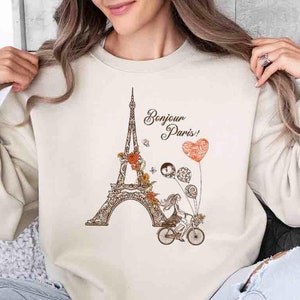 Pode incluir: Uma camisa de moletom bege com um gr&aacute;fico da Torre Eiffel, uma mulher andando de bicicleta e o texto "Bonjour Paris!" em letras marrons.