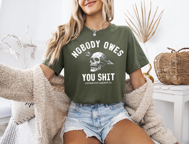 May include: An olive green t-shirt featuring the white text "NOBODY OWES YOU SHIT" and a skull graphic. The shirt is from Combat Iron Apparel Co. and is worn with denim shorts.