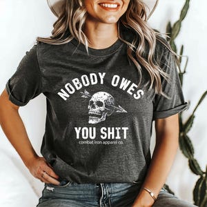 May include: Dark grey t-shirt with the text "NOBODY OWES YOU SHIT" in white, and a skull pierced by an arrow. The shirt also features the text "combat iron apparel co." below the main text.