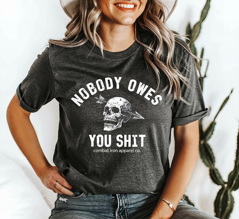 May include: Dark grey t-shirt with the text "NOBODY OWES YOU SHIT" in white, and a skull pierced by an arrow. The shirt also features the text "combat iron apparel co." below the main text.