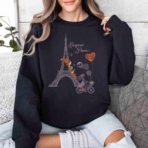 Pode incluir: Camisola de cor preta com um gr&aacute;fico da Torre Eiffel, um esqueleto a andar de bicicleta e o texto "Bonjour Paris!" em rosa.
