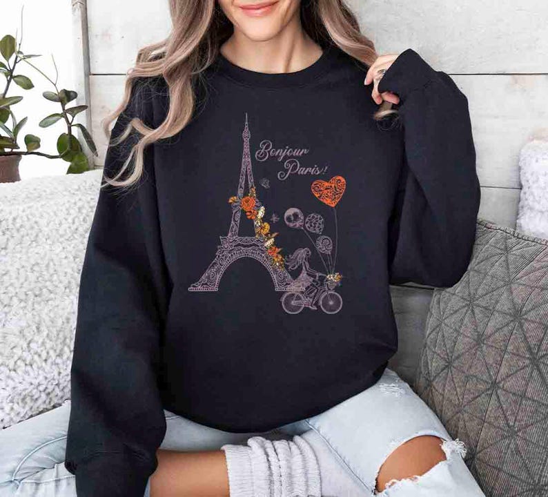 Pode incluir: Camisola de cor preta com um gr&aacute;fico da Torre Eiffel, um esqueleto a andar de bicicleta e o texto "Bonjour Paris!" em rosa.