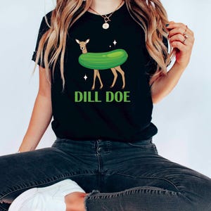 Puede incluir: Camiseta negra con un ciervo verde en forma de pepinillo y el texto "DILL DOE" en letras verdes.