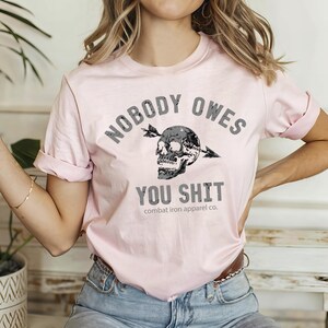 May include: A pale pink t-shirt featuring the slogan "NOBODY OWES YOU SHIT" in grey. The design includes a skull pierced by an arrow. The shirt is from "combat iron apparel co."