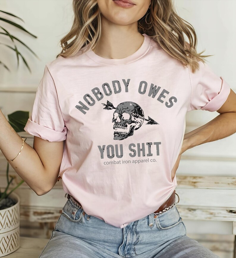 May include: A pale pink t-shirt featuring the slogan "NOBODY OWES YOU SHIT" in grey. The design includes a skull pierced by an arrow. The shirt is from "combat iron apparel co."