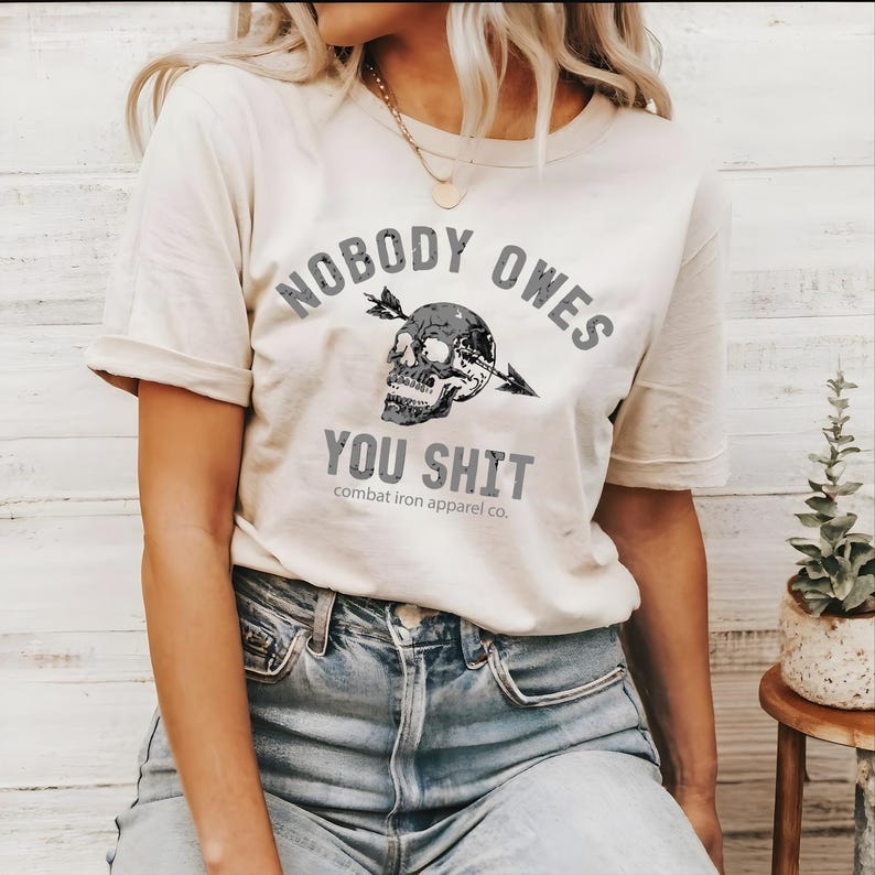May include: A cream-coloured t-shirt featuring the grey text "NOBODY OWES YOU SHIT" and a skull pierced by an arrow. The shirt is from "combat iron apparel co."