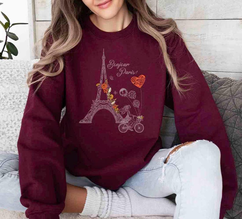 Pode incluir: Uma camisa de frio vinho com um gr&aacute;fico branco da Torre Eiffel, flores e uma pessoa andando de bicicleta. O texto "Bonjour Paris!" est&aacute; impresso na camisa de frio.