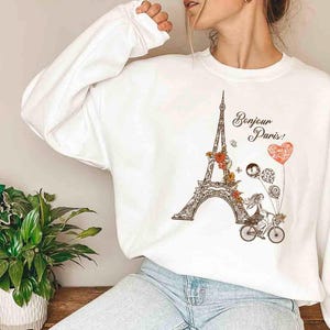 Pode incluir: Camisola branca com uma estampa gr&aacute;fica da Torre Eiffel, uma mulher de bicicleta e o texto "Bonjour Paris!" em letras pretas.