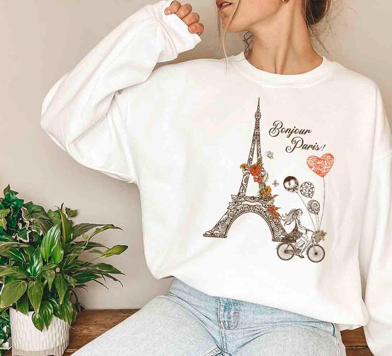 Pode incluir: Camisola branca com uma estampa gr&aacute;fica da Torre Eiffel, uma mulher de bicicleta e o texto "Bonjour Paris!" em letras pretas.
