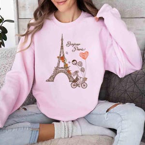 Pode incluir: Uma camisa de moletom rosa com um gr&aacute;fico da Torre Eiffel, uma mulher andando de bicicleta e o texto "Bonjour Paris!" em letras marrons.