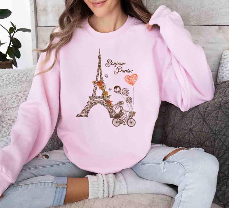 Pode incluir: Uma camisa de moletom rosa com um gr&aacute;fico da Torre Eiffel, uma mulher andando de bicicleta e o texto "Bonjour Paris!" em letras marrons.