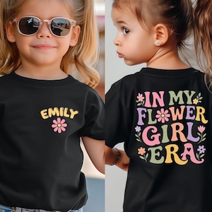 May include: Black t-shirt with the name "Emily" and a pink flower graphic on the front. The back of the shirt says "In My Flower Girl Era" in colorful, retro-style lettering. The shirt is worn by a young child.
